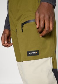 Calças de exterior verde-oliva com um bolso com fecho preto, secção inferior bege e um patch com o logótipo "IC3PARK" na coxa.