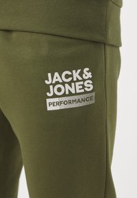 Grüne Baumwoll-Sweatpants mit geribtem Bund, versehen mit weißem Text "JACK & JONES PERFORMANCE" auf dem Oberschenkel. Glatte Textur.