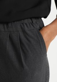 Pantalon gris foncé avec taille élastique, doté de poches latérales et d'une texture de tissu subtile. La coupe est ample avec des plis décoratifs.