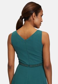 Zöld, ujjatlan ruha V-hátkivágással, díszes gyöngyökkel ellátott övvel és rejtett cipzárral a háton. Simább szövettextúra.