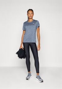 Sporty antrekk med en blå heather T-skjorte og svarte leggings. Modellen holder et svart håndkle. Grå joggesko med teksturert design.
