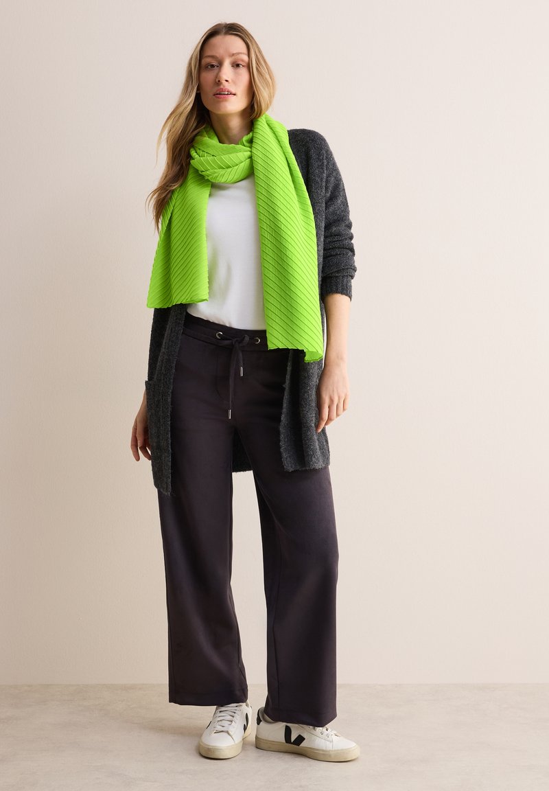 Un cardigan gris par-dessus un t-shirt blanc, associé à un pantalon large noir et une écharpe verte texturée brillante. Des baskets blanches avec des accents noirs.