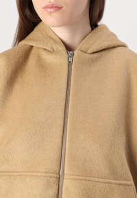 Beige hettejakke med glidelås laget av mykt stoff med en glatt tekstur. Den har en sølvfarget glidelås og et enkelt, minimalistisk design.