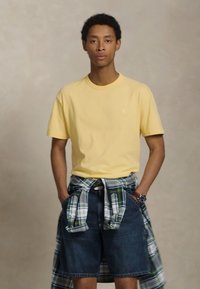 Ung mann iført en gul T-skjorte, rutete skjorte knyttet rundt livet, denimshorts og et armbåndsur, står med hendene i lommene.