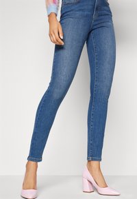 Vaqueros ajustados de mezclilla azul con cintura de tiro medio, con un corte slim y un sutil detalle de desgaste. Combinados con zapatos de tacón alto de color rosa claro.