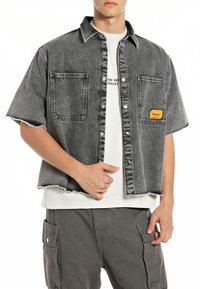 Camicia a maniche corte in denim grigio con orlo sfilacciato, dotata di due taschini sul petto e un'etichetta gialla. Indossata sopra una maglietta bianca.