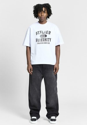 T-shirt blanc oversize avec texte noir "ATELIER 2018 RETERNITY DIRECTEUR CRÉATIF," associé à un jean large sombre et des baskets noires.