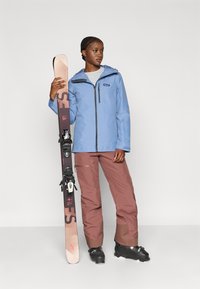 Ανοιχτό μπλε αδιάβροχο μπουφάν με φερμουάρ μπροστά, γκρι πουκάμισο από κάτω, καφέ παντελόνι σκι και κρατάει ένα σκι με μαύρες δεσίματα.