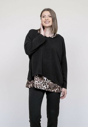 Femme souriante portant un pull noir ample superposé à un chemisier imprimé léopard et un pantalon noir, debout devant un fond clair uni.