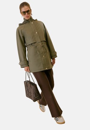 Frau trägt grüne Kapuzenjacke, braune weite Hose, weiße Sneakers, Sonnenbrille und trägt eine dunkel geflochtene Tragetasche.