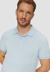 Jasnoniebieska koszulka polo z teksturowanej tkaniny, posiadająca kołnierzyk, zapięcie na trzy guziki oraz krótkie rękawy z mankietami. Prosty design bez wzorów.