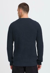 Man with short hair and hoop earring wearing a navy blue ribbed knit sweater, facing away against a plain light background.