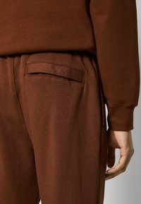 Detailní záběr na hnědé tepláky s kapsou na zadní straně, nošené s odpovídajícím hnědým dlouhým rukávem, ukazující uvolněný střih a měkkou látku.