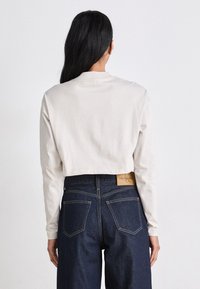 Chemise beige à manches longues raccourcie avec un col rond, associée à un jean en denim foncé taille haute avec un patch de marque brun et deux poches arrière.