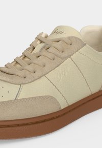 Baskets en cuir beige et crème avec accents en daim, lacets beiges, logo en relief sur le côté et semelle en caoutchouc marron visible sur fond blanc.