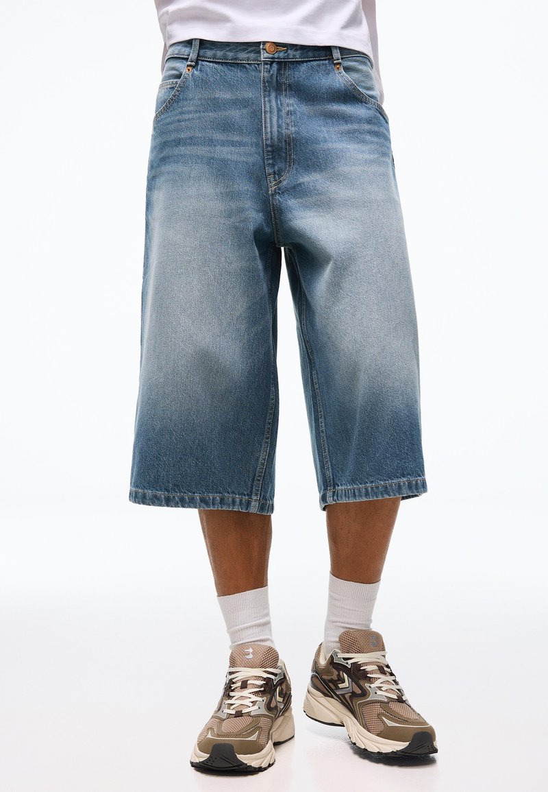 Homme portant un short en jean bleu clair mi-genoux, des chaussettes blanches et des baskets beiges, debout devant un fond uni.