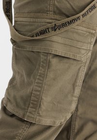 Pantalones cargo verde oliva hechos de tela duradera, con múltiples bolsillos, detalles de costura y una etiqueta con el texto "QUITAR ANTES DEL VUELO".