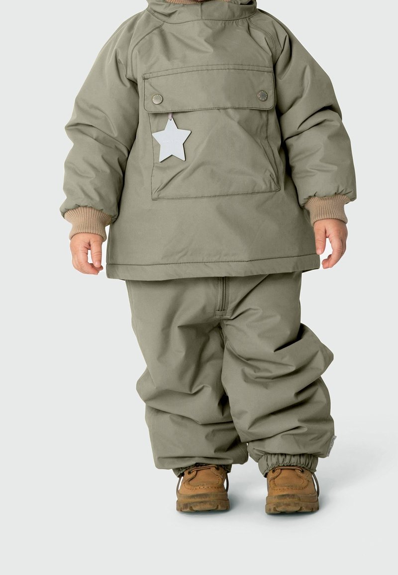 Peuter draagt een olijfgroen gewatteerd jasje en broek, met bruine laarsjes en een stervormig reflecterend label op de zak van het jasje.
