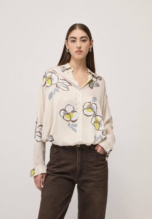 Femme aux longs cheveux bruns portant un chemisier crème à boutons avec de grands motifs floraux et un jean foncé taille haute, debout devant un fond uni.