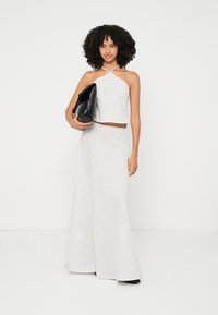 Top corto blanco estilo halter y pantalones de pierna ancha con rayas verticales negras; la modelo sostiene una bolsa negra grande, tela texturizada, diseño minimalista.