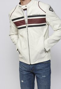 Giacca bianca con strisce orizzontali nere e marroni, chiusura frontale con zip, toppa sulla manica sinistra, indossata con jeans blu strappati, mani nelle tasche.