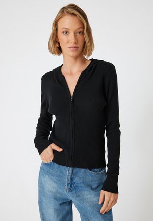 Zwarte geribbelde zip-up hoodie met een aansluitend ontwerp, lange mouwen en een capuchon, gedragen over losse denim jeans. De stof lijkt gestructureerd.