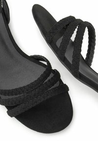 Schwarze Sandalen mit geflochtenen Riemen, aus weichem Wildleder gefertigt. Die Sohle ist flach, mit einer glatten Textur und einem offenen Zehenbereich.