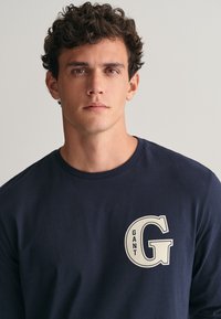 Námořnická dlouhá trička vyrobená z měkkého materiálu, s krémovým logem "GANT" a velkým "G" na levé části hrudi. Jednoduchý design.