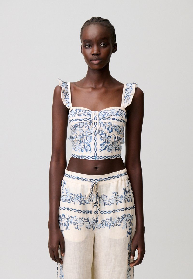 Giovane donna che indossa un completo crop top color crema e pantaloni con ricamo floreale blu, in piedi davanti a uno sfondo grigio chiaro uniforme.