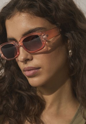 Mujer con el cabello largo y rizado, que lleva gafas de sol rectangulares translúcidas rosas y pendientes de aro plateados, mirando ligeramente hacia abajo.