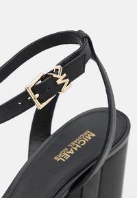 Sandale à talon haut en cuir noir avec boucle dorée et logo « MK » sur la bride de cheville, nom de la marque Michael Kors sur la semelle intérieure.