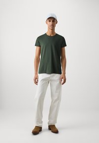 Grünes Baumwoll-T-Shirt, weiße Jeanshose und braune Wildlederstiefel. Hellblaues Cap. Einfaches Design mit lockerer Passform. Einfache Farbe ohne Muster.