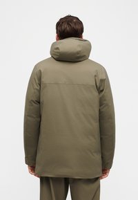 Άνδρας που φοράει έναν χειμερινό παλτό με κουκούλα σε ελιά πράσινο, γυρισμένος με την πλάτη, δείχνοντας τις λεπτομέρειες της πλάτης και τη στενή γραμμή του ενδύματος.