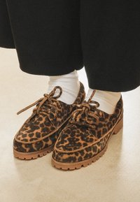 Scarpe in suede con stampa leopardata e dettagli marroni, punta rotonda, design con lacci e suola in gomma spessa, abbinate a calzini bianchi e pantaloni neri.