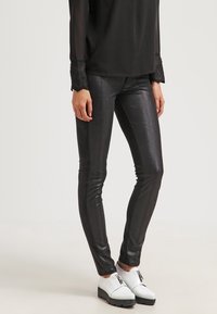 Zwarte, glanzende, strakke legging van een metallic materiaal, gecombineerd met witte schoenen met een ritssluiting en een dikke zool.
