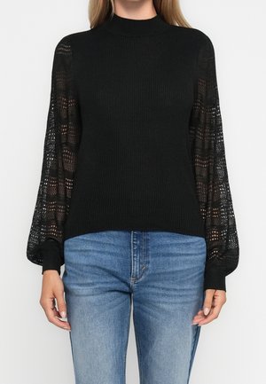 Sort ribstrikket sweater med gennemsigtige, mønstrede lange ærmer, stylet med blå denimjeans på en stående person.