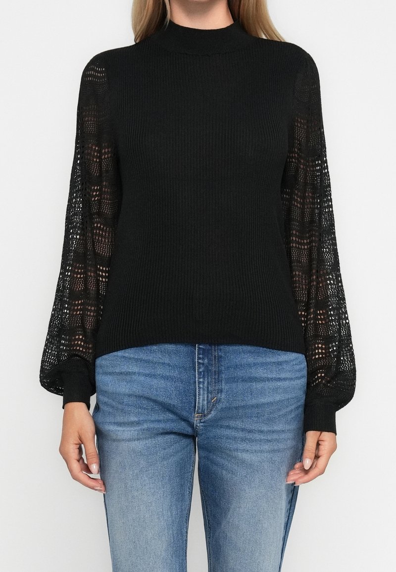 Pull noir côtelé avec manches longues transparentes à motifs, associé à un jean en denim bleu porté par une personne debout.