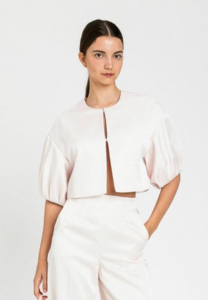 Donna con capelli scuri raccolti dietro che indossa un top corto bianco con maniche a sbuffo e pantaloni a vita alta abbinati su sfondo bianco.