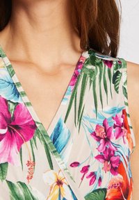 Floral sleeveless top featuring a v-neck design. Made of lightweight fabric, it showcases colorful tropical flowers and green leaves on a beige background.