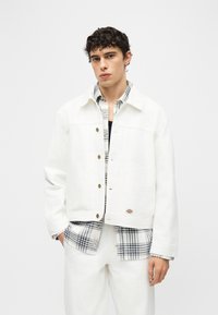 Νέος άνδρας με σγουρά μαλλιά που φοράει λευκό σακάκι με κουμπιά πάνω από ένα ασπρόμαυρο καρό πουκάμισο και λευκό παντελόνι μπροστά σε απλό φόντο.