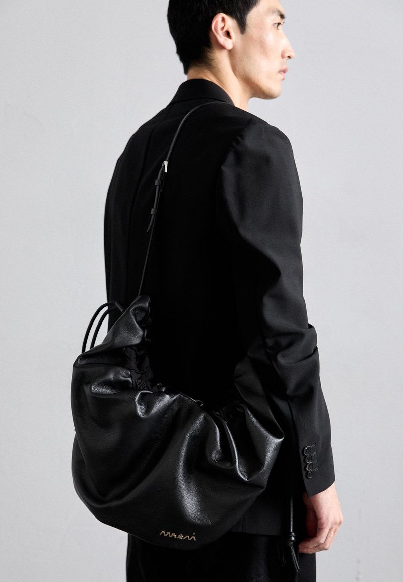 Uomo che indossa un blazer nero e porta una grande borsa a tracolla in pelle nera plissettata con una sottile catena, su uno sfondo grigio uniforme.