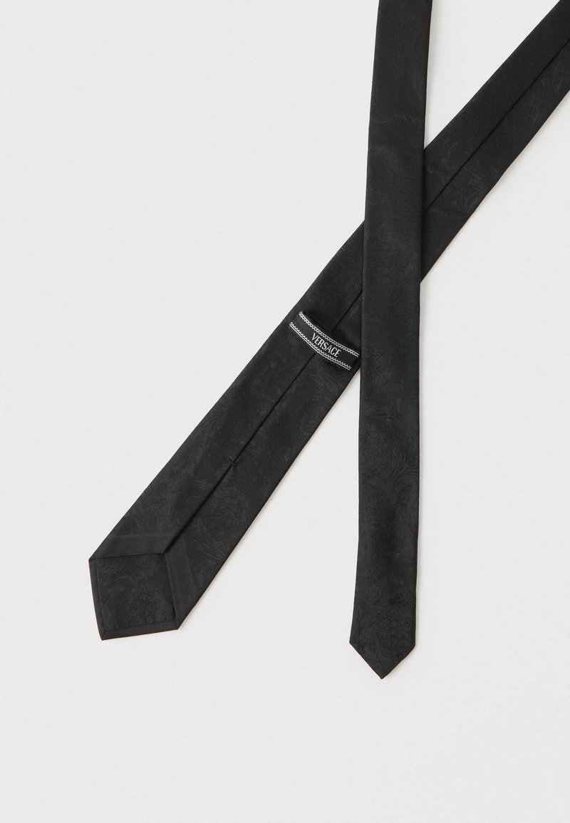 Cravate en soie noire avec un motif floral subtil, une forme effilée et une extrémité pointue. Comprend une étiquette de marque près de l'extrémité large.