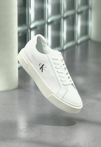 Λευκό χαμηλό αθλητικό παπούτσι Calvin Klein με κορδόνια, που αιωρείται πάνω από γυαλισμένο τσιμεντένιο δάπεδο σε φόντο από ματ γυάλινα τούβλα.