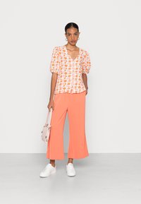 Blusa floral com mangas bufantes em laranja e rosa, combinada com calças largas coral e ténis brancos; a segurar uma bolsa rosa clara.