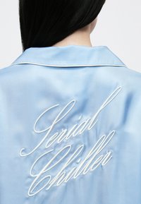 Világoskék szatén ing hegyes gallérral, fehér hímzett "Serial Chiller" felirattal a háton. Puha tapintású és fényes felületű.