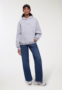 Felpa grigia chiara con tasca frontale e cappuccio con cordino, abbinata a pantaloni in denim blu a gamba larga e sneakers metallizzati. Texture del tessuto liscia.