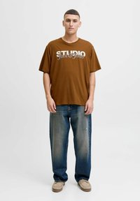 Brązowy bawełniany t-shirt z napisem "STUDIO Jack & Jones", w zestawieniu z wyblakłymi niebieskimi dżinsami i beżowymi zamszowymi butami. Luźny krój, krótki rękaw.