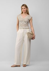 Beige Oberteil im Chevron-Muster mit kurzen Ärmeln, kombiniert mit cremefarbenen, weit geschnittenen Hosen. Hält eine beigefarbene Handtasche mit Kettenriemen.