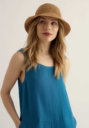 Kobieta o długich blond włosach, ubrana w niebieski top bez rękawów i brązowy pleciony kapelusz przeciwsłoneczny, patrząca nieco z boku z neutralnym wyrazem twarzy.