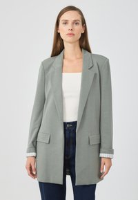 Blazer ajusté gris clair avec des revers, doté de deux poches avant et de poignets retroussés, sur un haut blanc, assorti à un jean foncé taille haute.